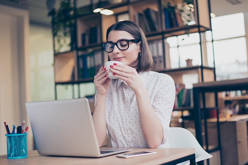 a white woman sitting at a desk with a laptop and cup of coffee