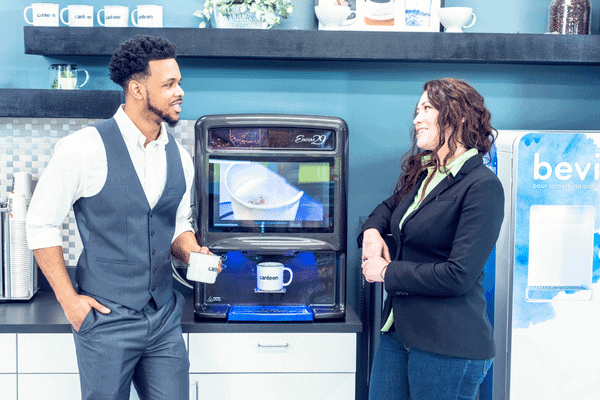 a black man and a white woman stand in front of a coffee machine